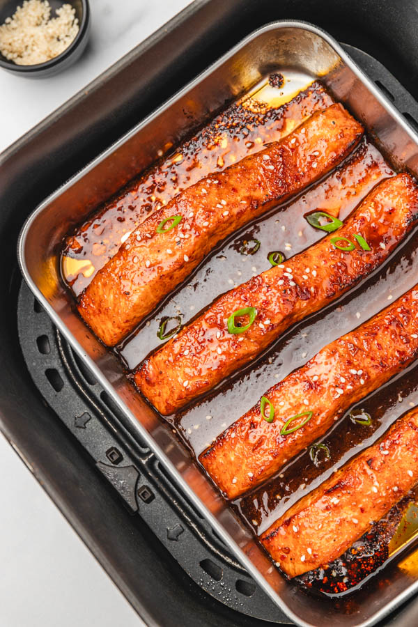 four salmon fillets in an air fryer basket garnished with chopped green onions and sesame seeds.