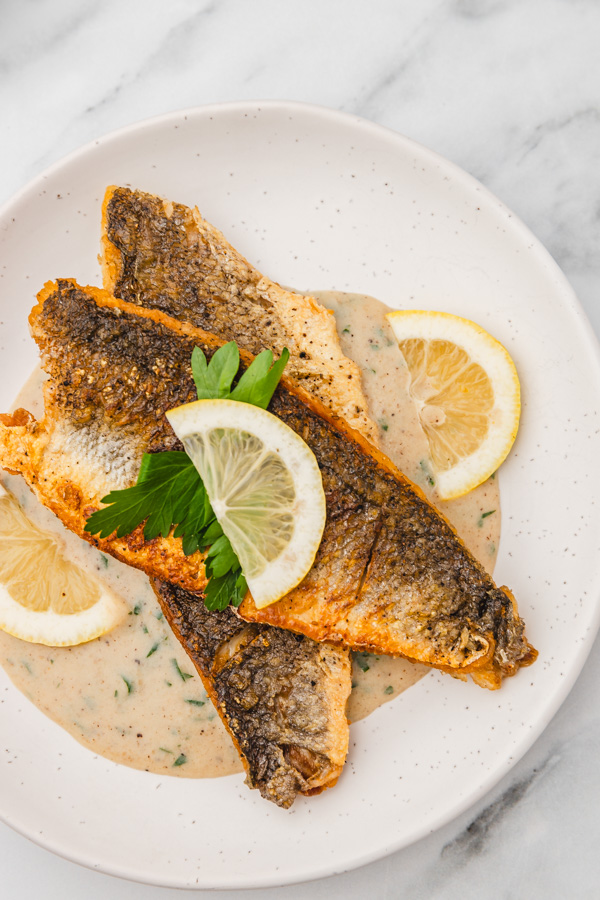 a plate of two pan fried sea bass fillets garnished with lemon slices and fresh parsley on a bed pf creamy sauce.