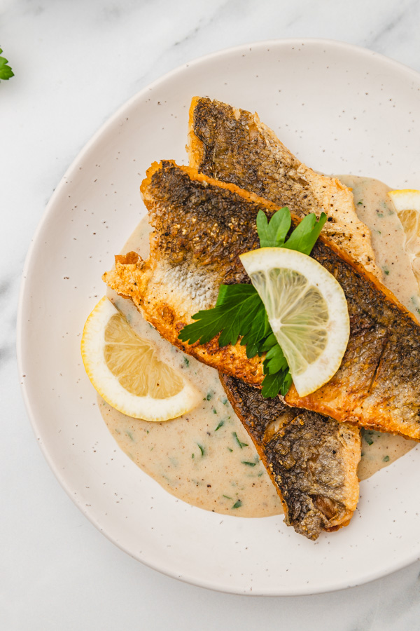 two pan seared sea bass fillets on a bed of creamy sauce. The sea bass is garnished with fresh parsley and lemon slices.