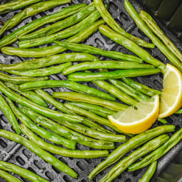 Green beans in air fryer basket with two lemon wedges.