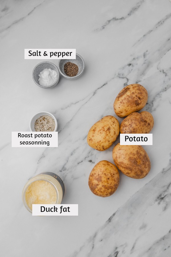 ingredients needed to make roast potatoes laid on a white marble surface.