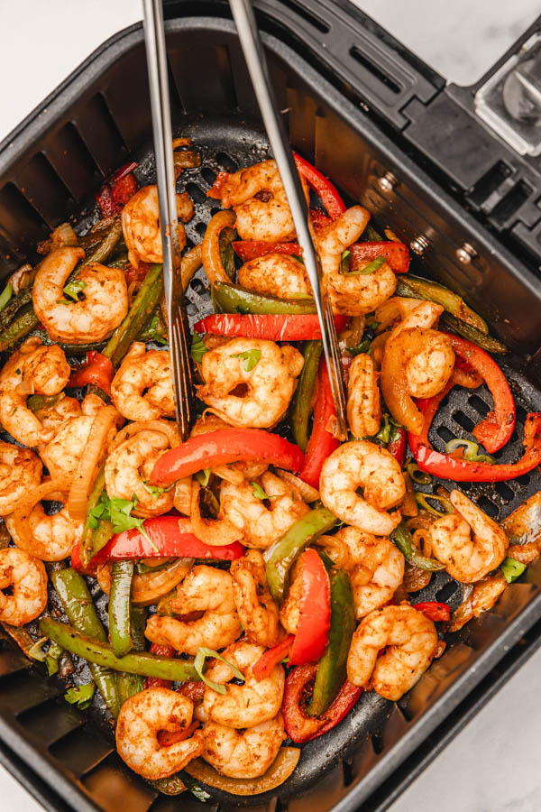 a metal tong in air fryer basket with shrimps and peppers. 