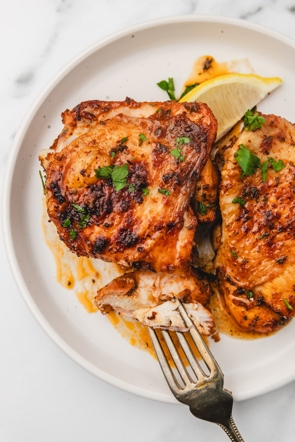 two chicken thighs on a plate with a fork and a lemon wedge.