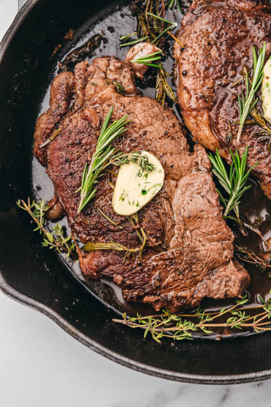 two perfectly cooked beef steak resting in a skillet topped with butter and fresh spring of rosemary and thyme.