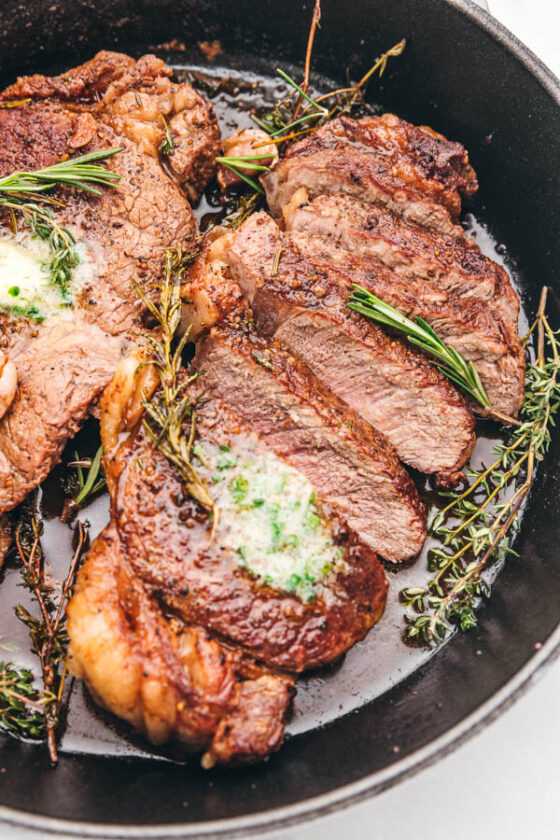 a close up of sliced thick steak in a skillet topped with melting herb butter and a spring of rosemary.
